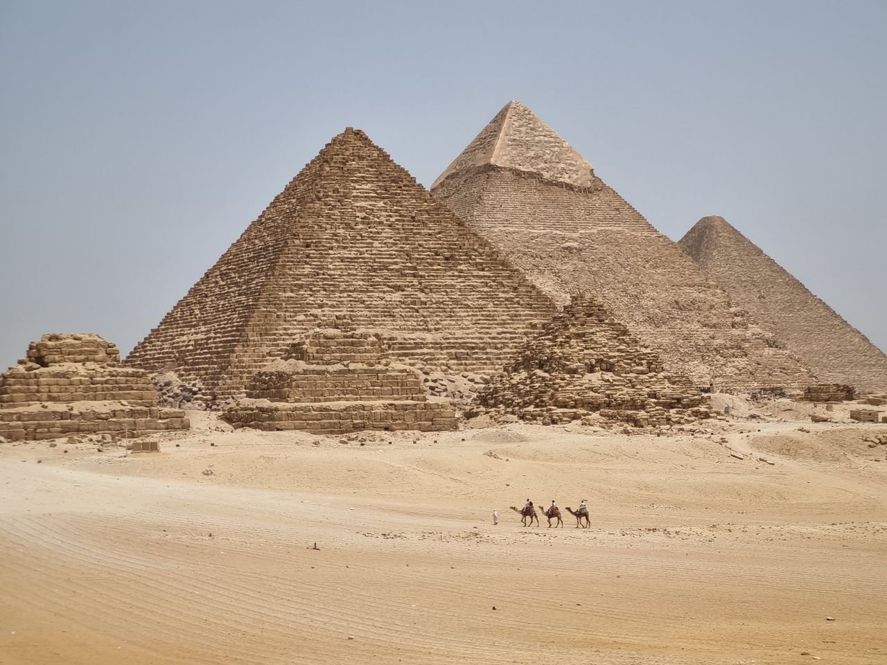 Panoramic shot of the Pyramids of Giza with camel riders in the foreground under clear skies.